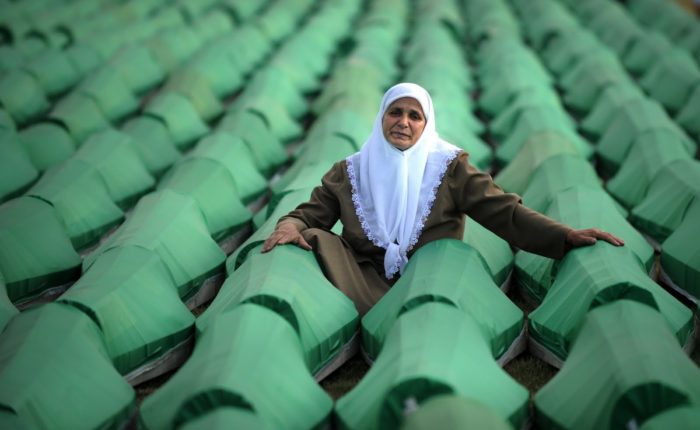 An elderly woman in a white headscarf sits among green coffins containing remains of Srebrenica massacre victims, awaiting burial.