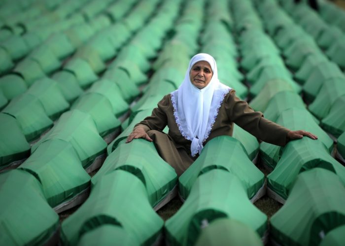 An elderly woman in a white headscarf sits among green coffins containing remains of Srebrenica massacre victims, awaiting burial.