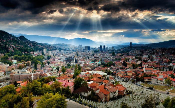 Panoramic view of Sarajevo city from a high vantage point, showcasing the city's dense architecture, surrounding mountains, and dramatic clouds with sun rays breaking through.
