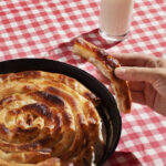 A freshly baked spiral-shaped burek in a cast iron pan, served with a glass of yogurt on a red-and-white checkered tablecloth.