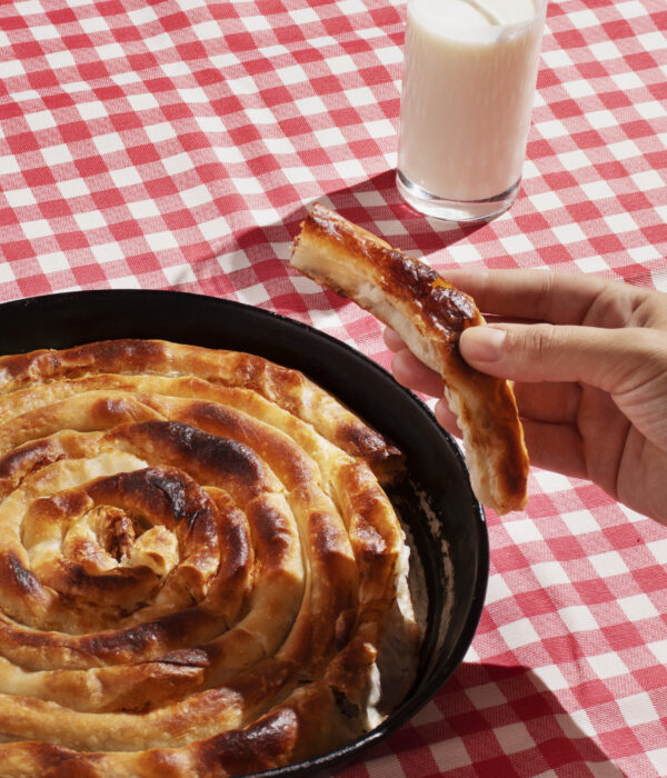 A freshly baked spiral-shaped burek in a cast iron pan, served with a glass of yogurt on a red-and-white checkered tablecloth.